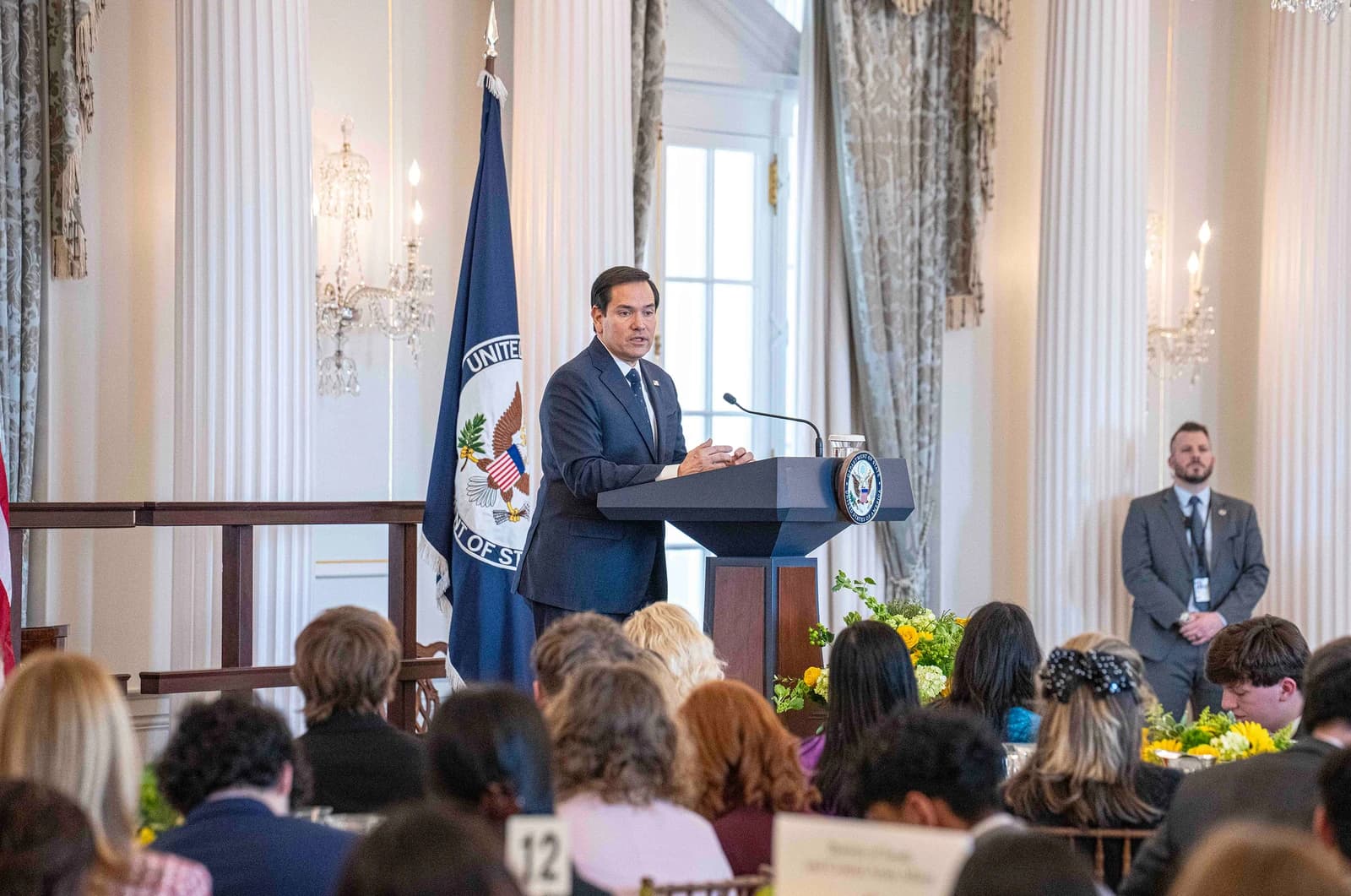 Senator Marco Rubio speaking at a podium to an audience in a formal room.