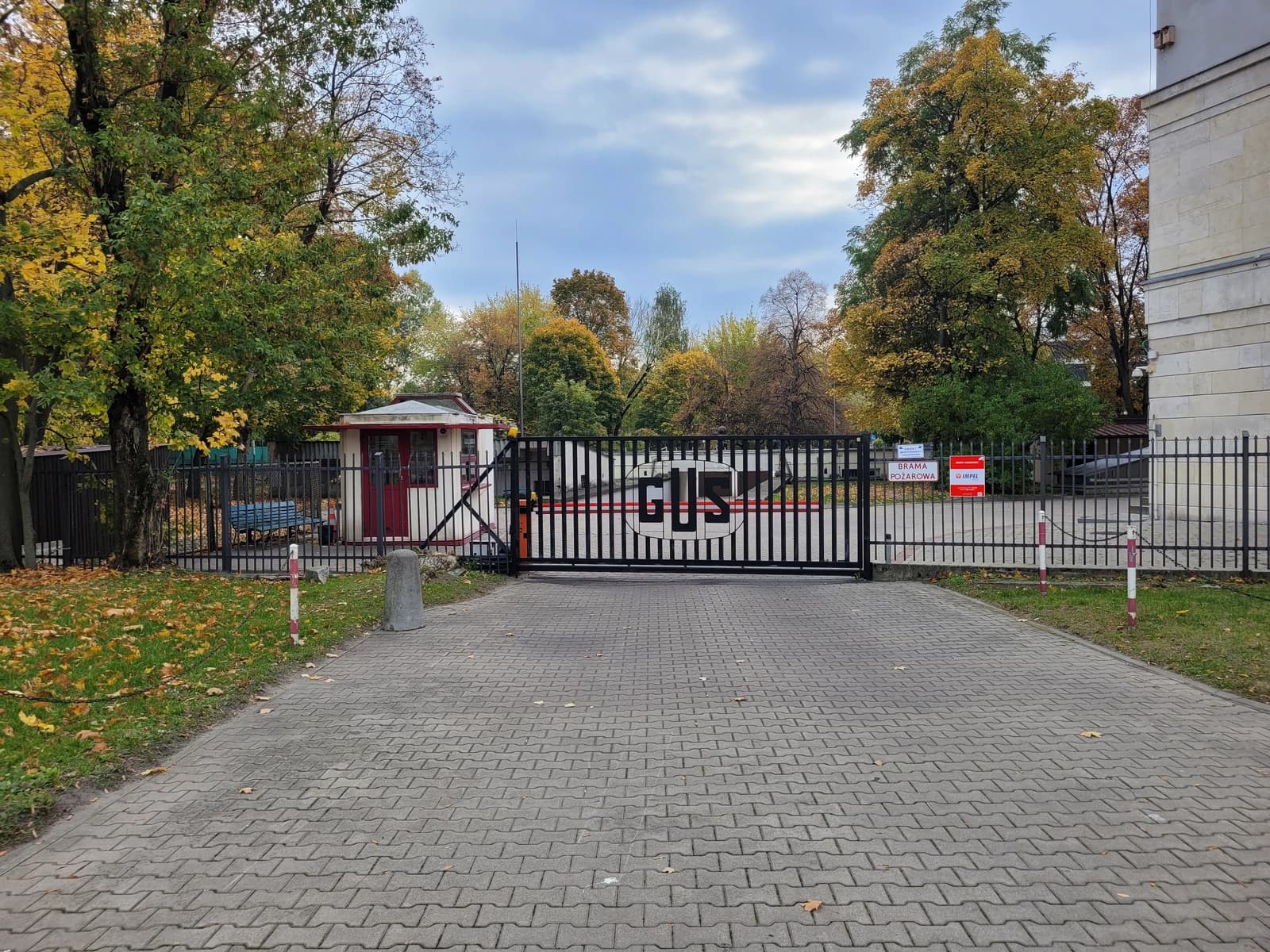 Entrance gate with "GUS" logo, small security booth, and trees in Warsaw.