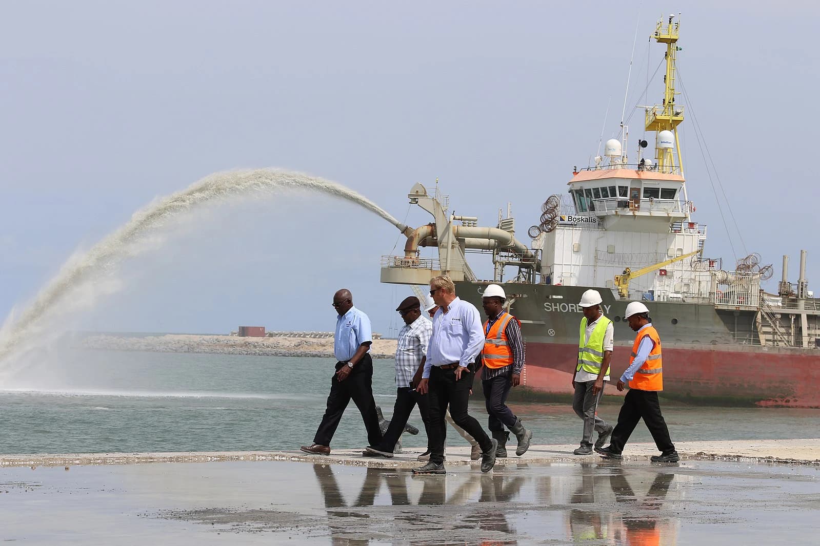 Men in various attire walking along a wet pier with a large vessel behind them.