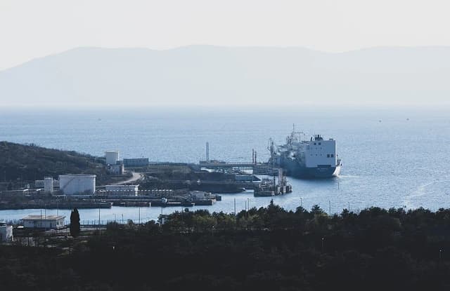 An LNG carrier ship docked at a port with industrial facilities along the coastline.