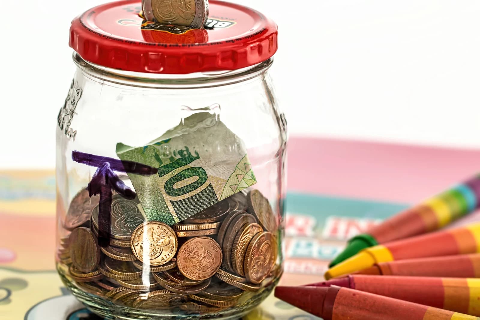 A clear glass jar filled with various coins and a folded ten rand banknote.