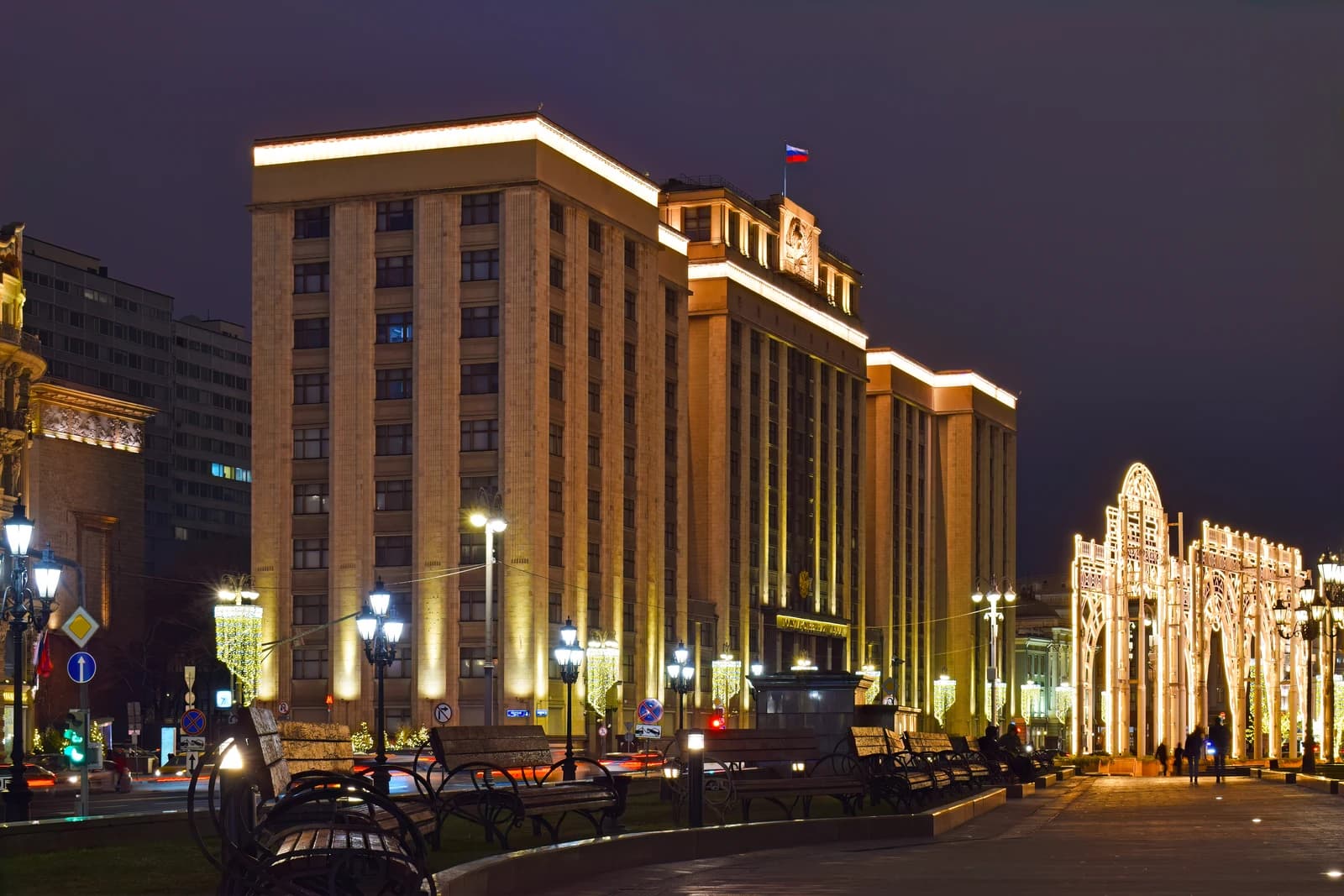 The State Duma building in Moscow at night, with Russian flag visible.