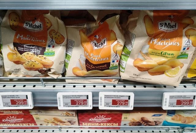 French madeleine cookies on a supermarket shelf with Cyrillic price tags.