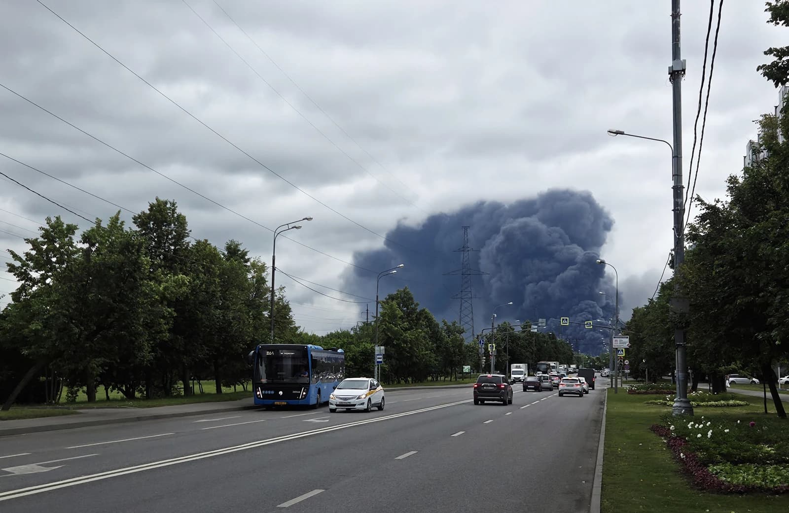 Thick black smoke plumes from a fire seen from a road with cars and a bus in Moscow.