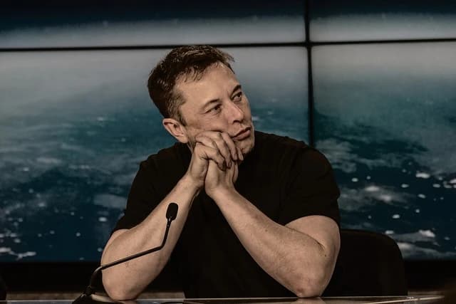 Elon Musk, CEO of Tesla, sitting at a table with hands clasped, looking away.