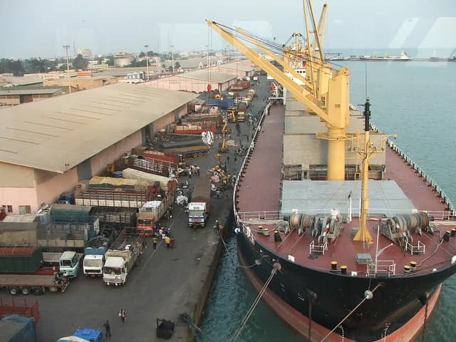 Cargo ship docked at the Port of Cotonou, Benin, with cranes and trucks.