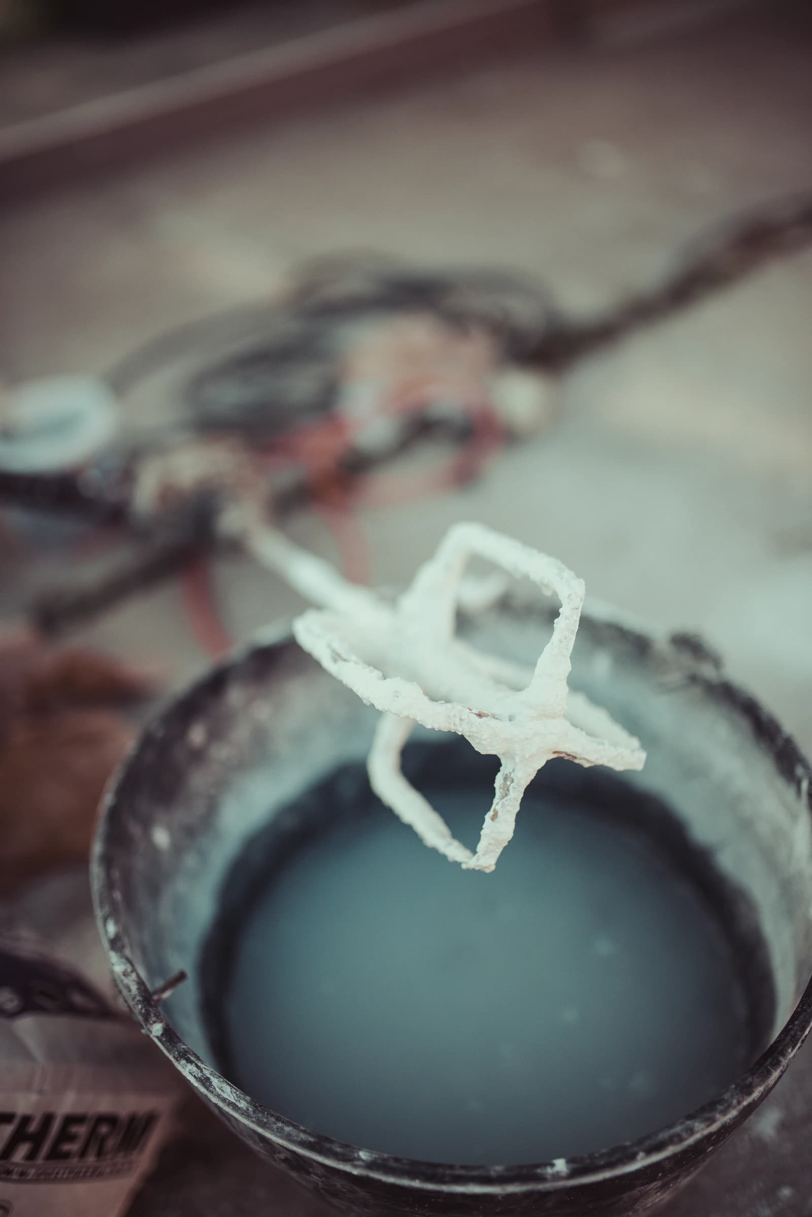 Construction mixing tool covered in dry materials, above a bucket of liquid at a job site.