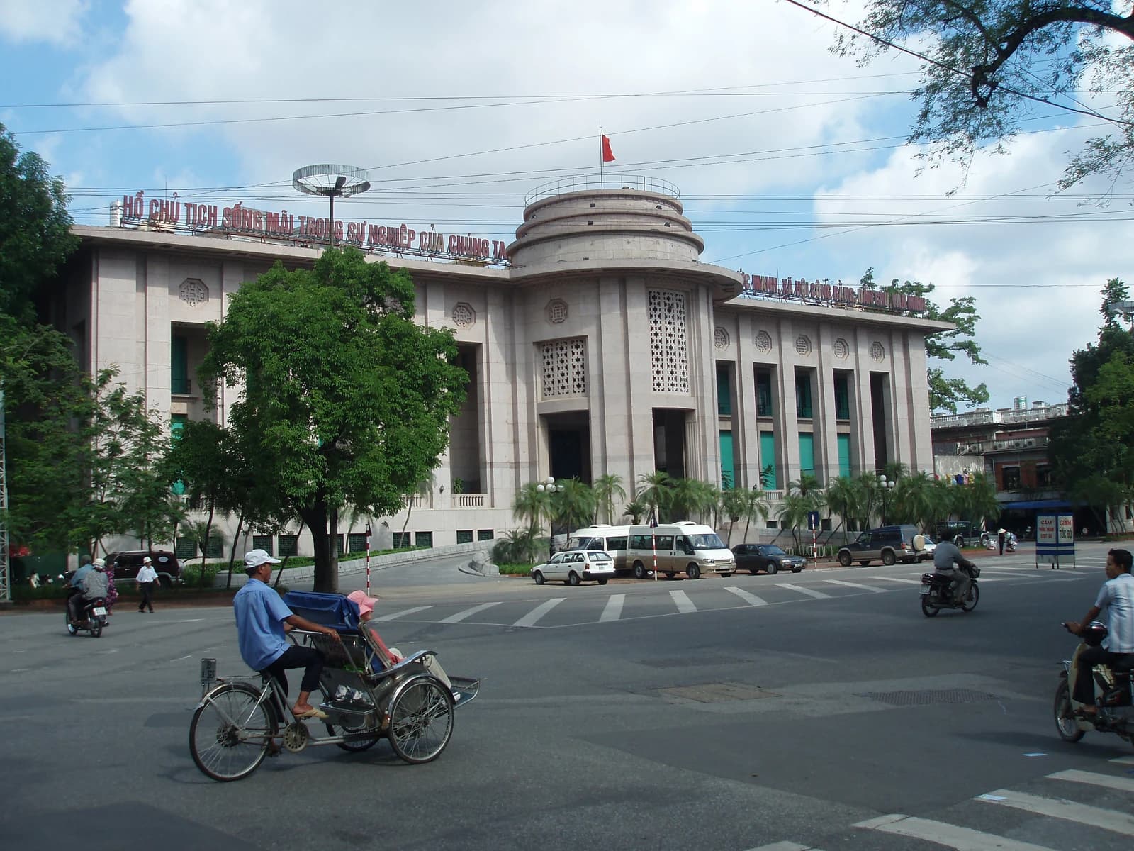 The State Bank of Vietnam headquarters building, a large stone structure with a circular roof section, stands on a street in Hanoi.