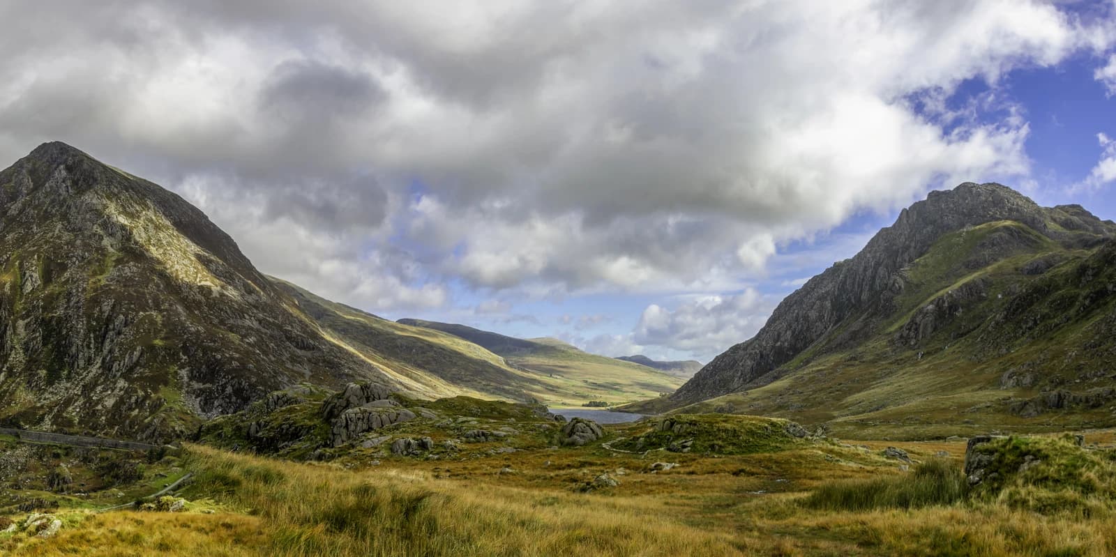 Panoramic view of a mountainous valley with a lake under a cloudy sky.