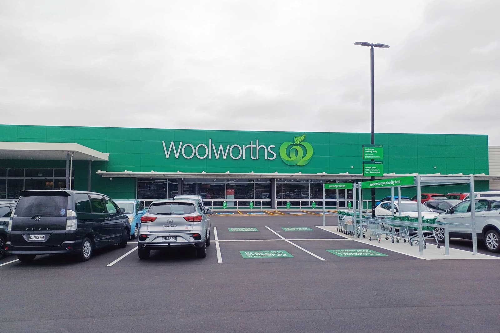 Exterior of Woolworths supermarket building with cars in the parking lot under a cloudy sky.