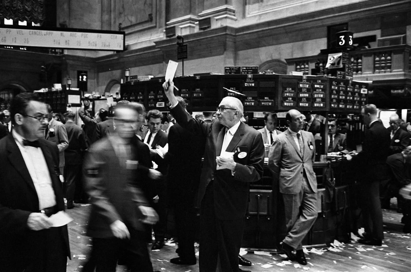 Traders in suits on the New York Stock Exchange floor, one raises a paper.