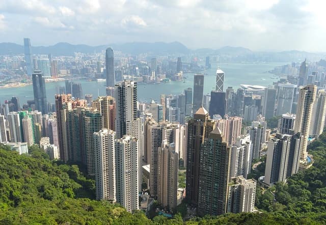 Hong Kong skyline with numerous skyscrapers, Victoria Harbour, and distant mountains.