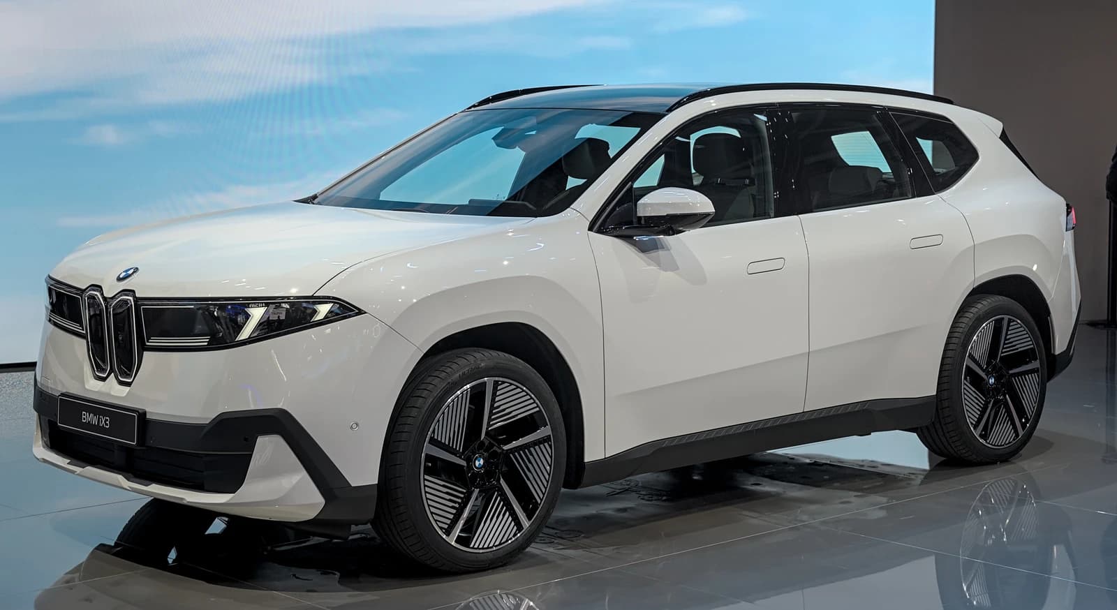 White BMW iX3 electric SUV displayed at an auto show with a light blue background.