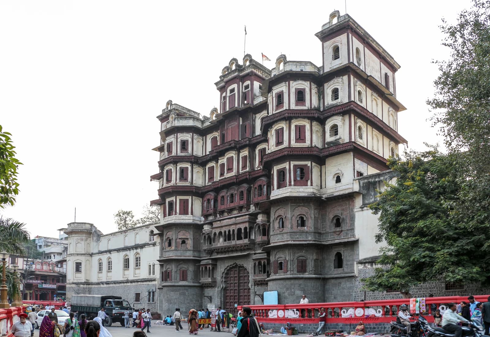People walking and vehicles parked on a street in front of Rajwada Palace in Indore, India.