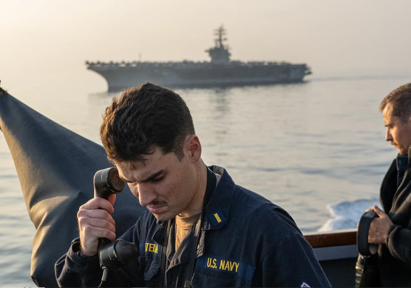 US Navy sailor holds communication device on deck; aircraft carrier USS Dwight D. Eisenhower visible.