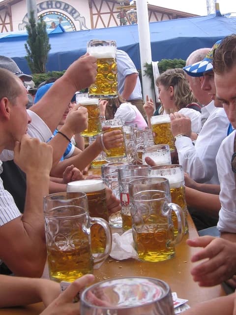 People at Oktoberfest holding large beer steins at a long wooden table.