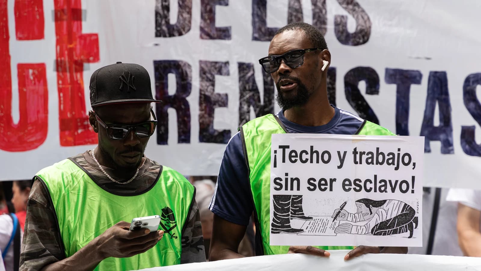 Two men demonstrating in Madrid with a sign that reads "¡Techo y trabajo, sin ser esclavo!".