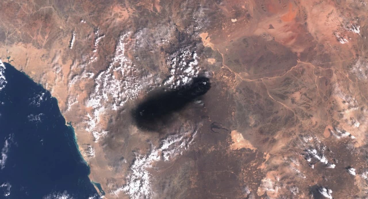 Satellite image showing a large dark smoke plume originating over land near a coastline.