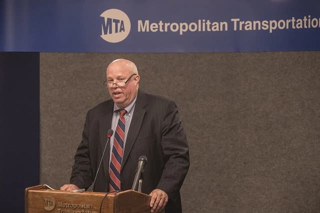 Man in suit and tie speaking at a podium with microphones at a press conference.