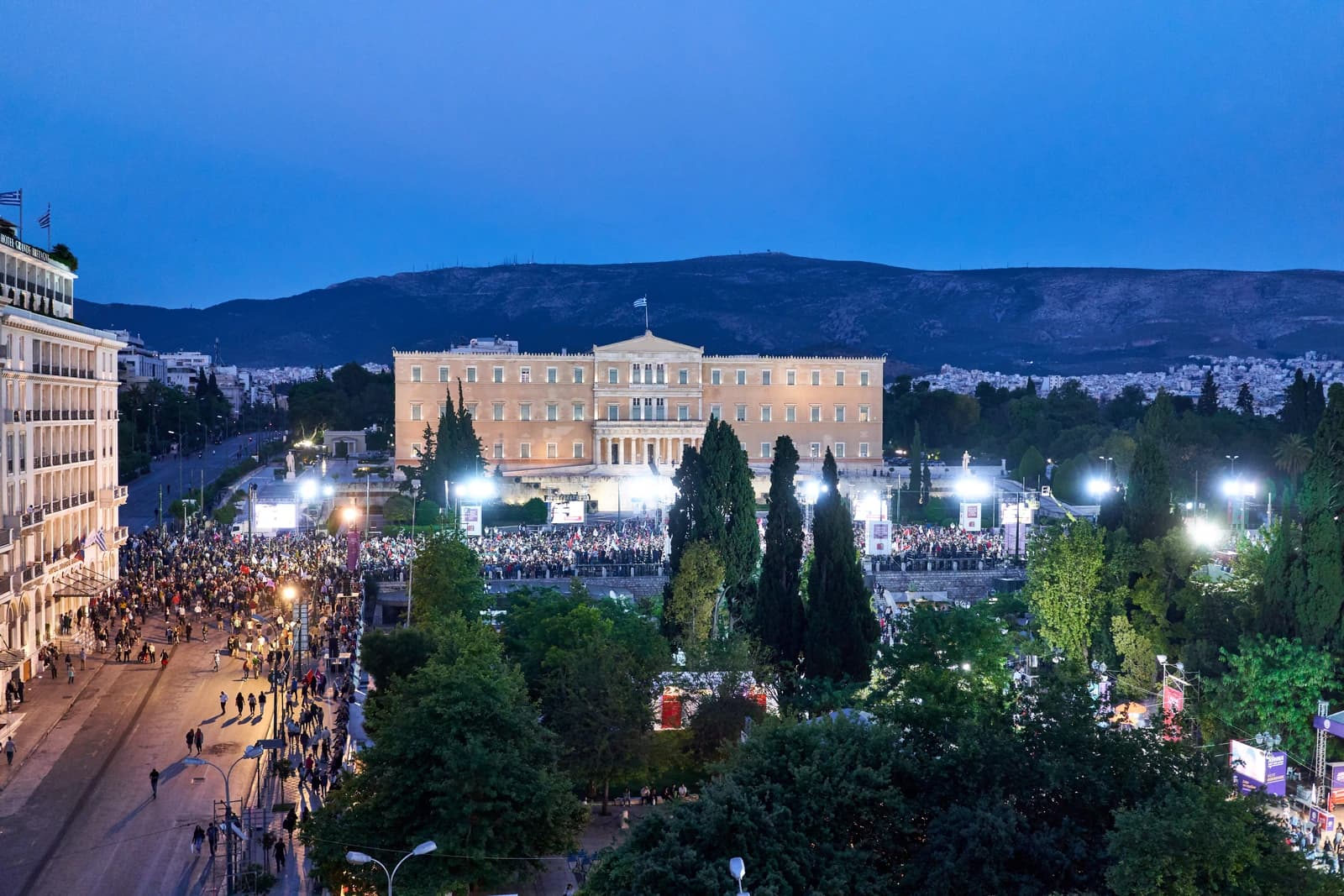 The Hellenic Parliament building illuminated at dusk with a crowd in Syntagma Square.