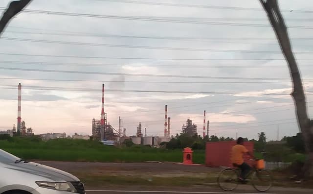 An oil refinery with multiple red and white striped smokestacks and storage tanks under a cloudy sky.