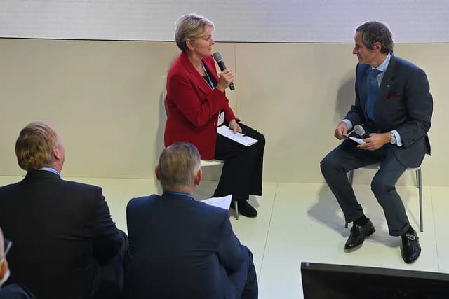 A woman in a red jacket speaks into a microphone while a man in a suit listens.