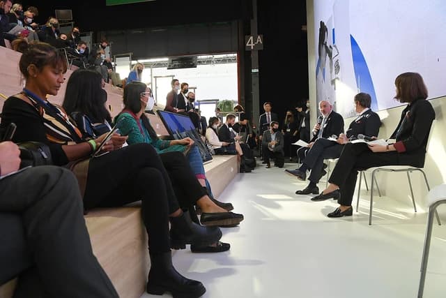 A male panelist speaks into a microphone while two female panelists listen at a conference.