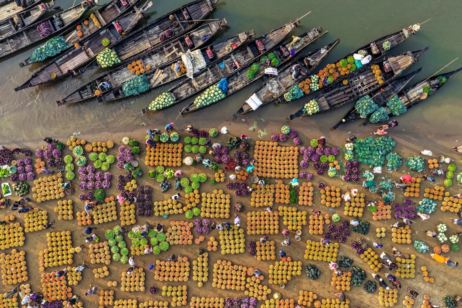 Aerial view of a floating market with people trading vegetables and fruits from boats and shore stalls.