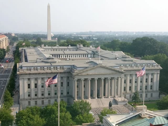 The United States Treasury Building stands with American flags and Washington Monument behind.
