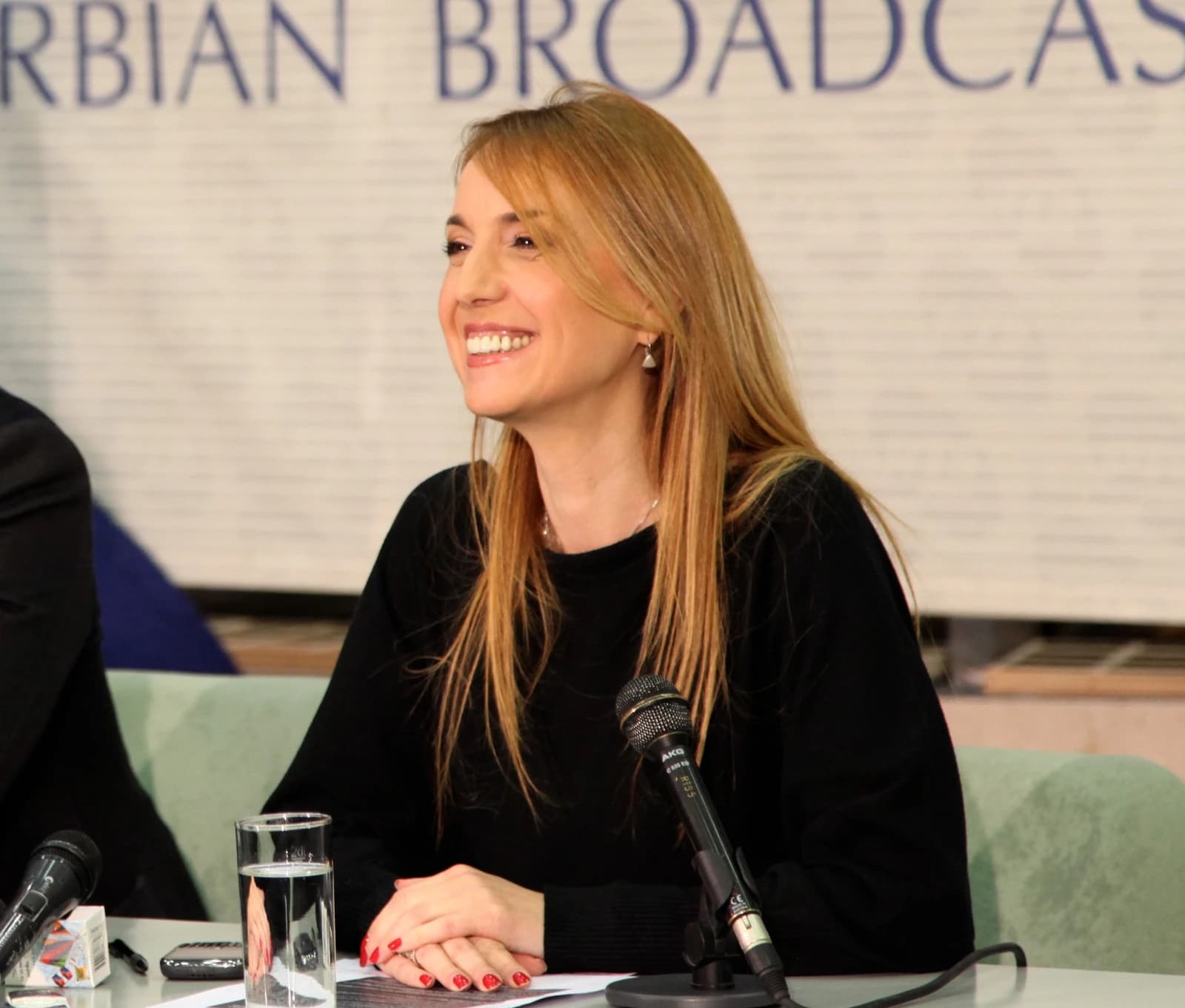Duška Vučinić, a TV presenter, smiling at a press conference table with a microphone.