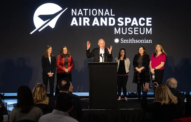 A man gestures while speaking at a podium with six women standing behind him.