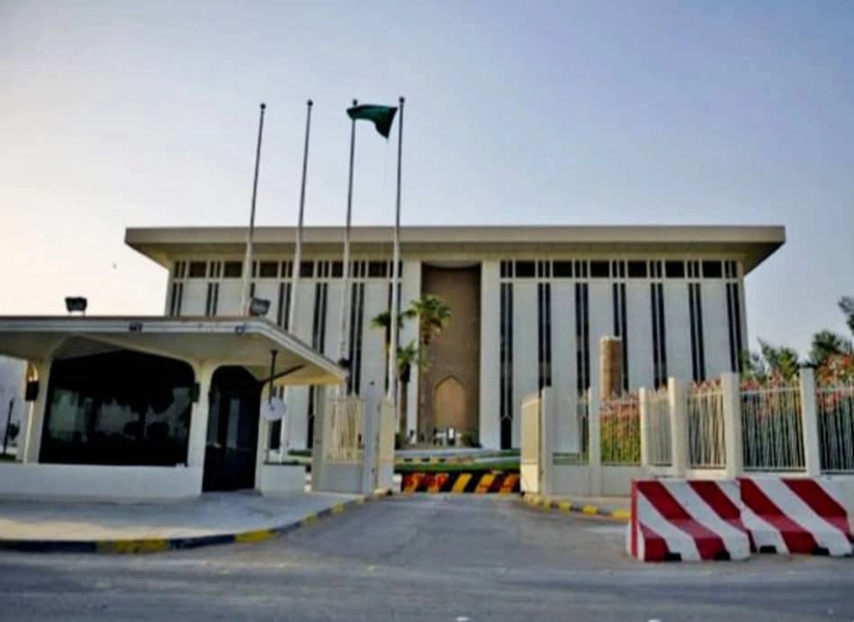 Exterior of a bank building with a Saudi flag flying and security booth in the foreground.