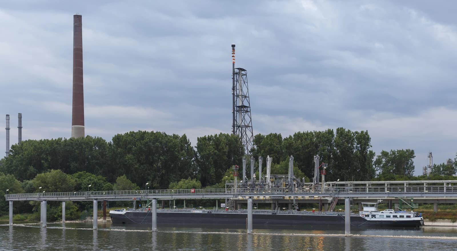 An oil refinery with a tall chimney, industrial structures, and a barge on a river.