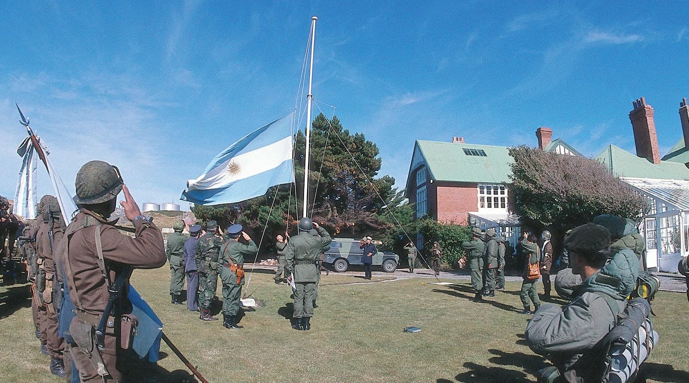 Argentine soldiers in uniform saluting an Argentine flag being raised on a flagpole.