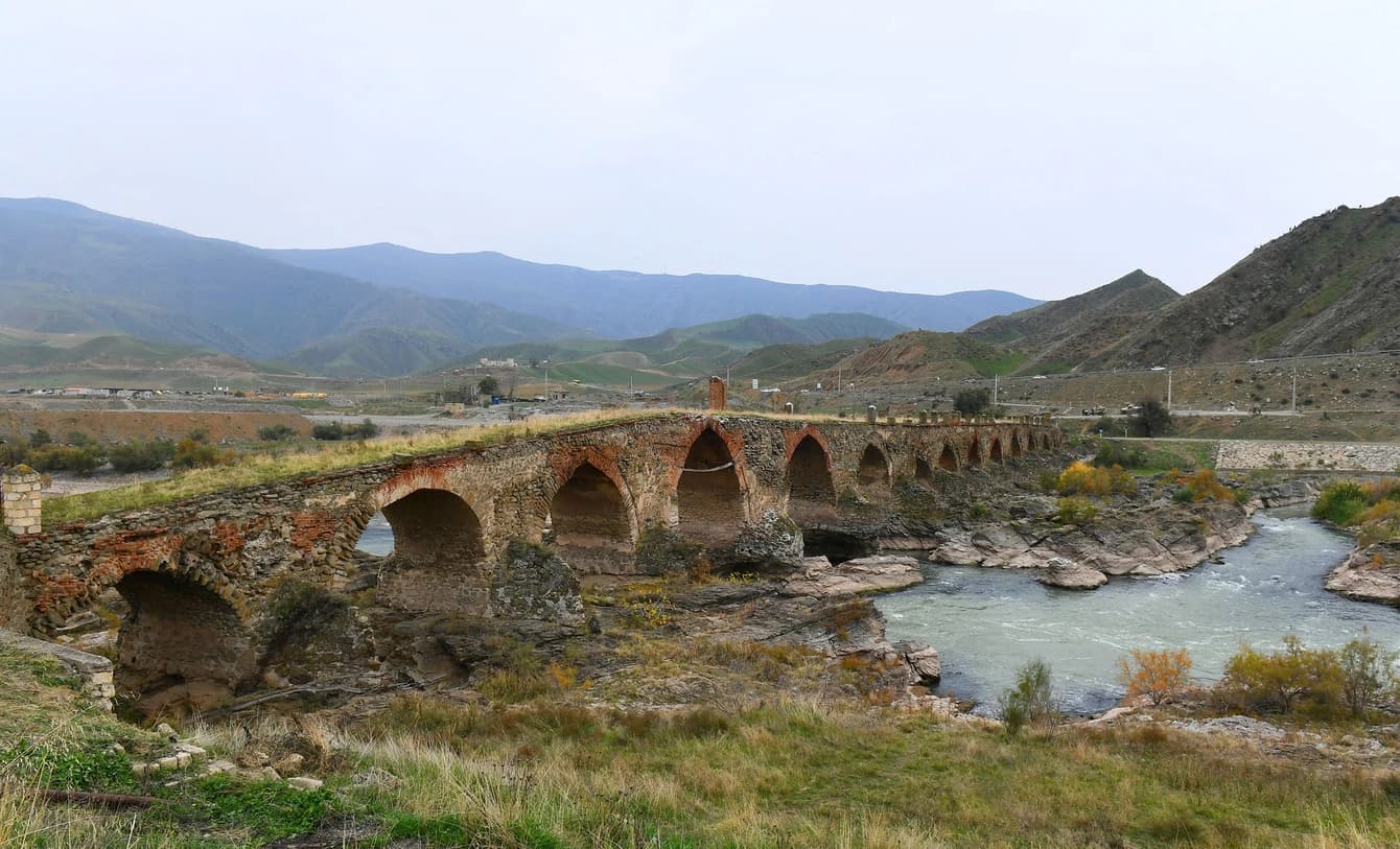Ancient Khudafarin Bridge, an arched stone structure, spans a river in a mountainous landscape.