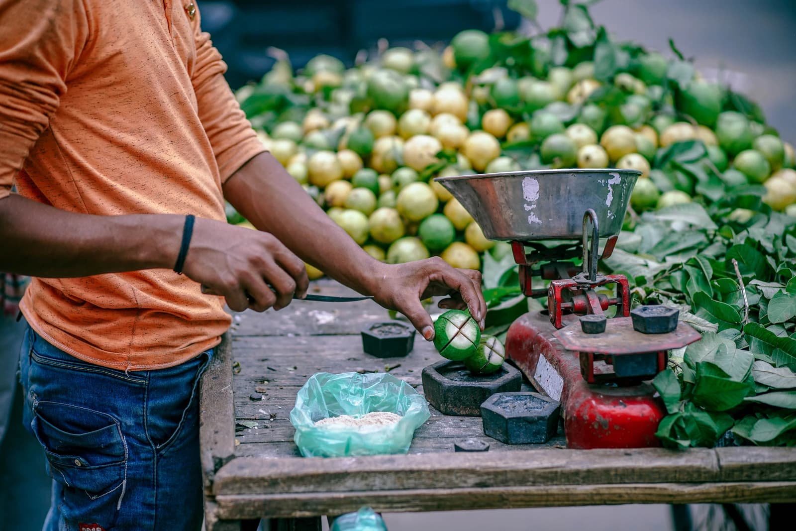 Street fruit seller cutting green fruit with a small knife next to a weighing scale.