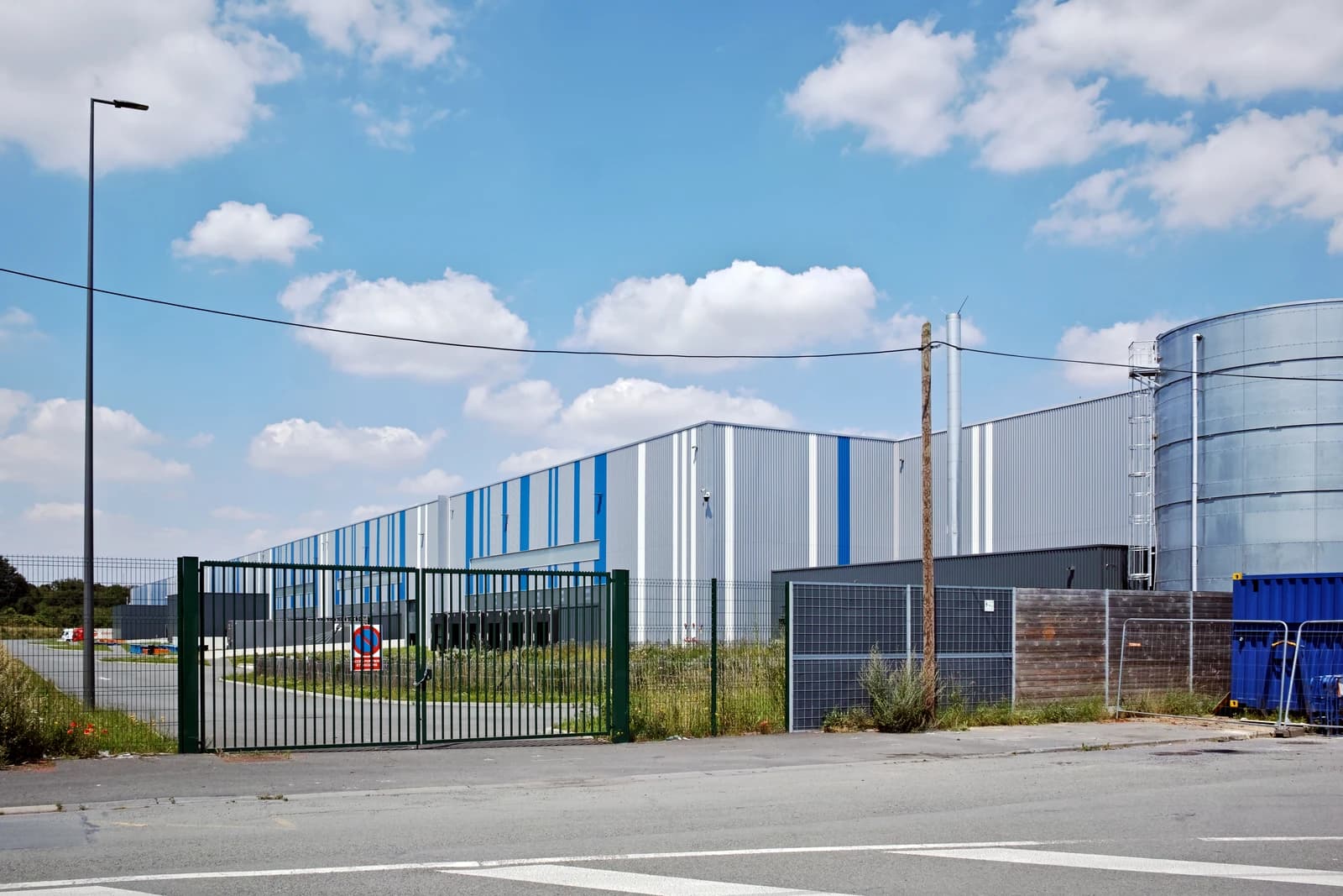 Exterior view of an industrial warehouse with a large fence and road in foreground.
