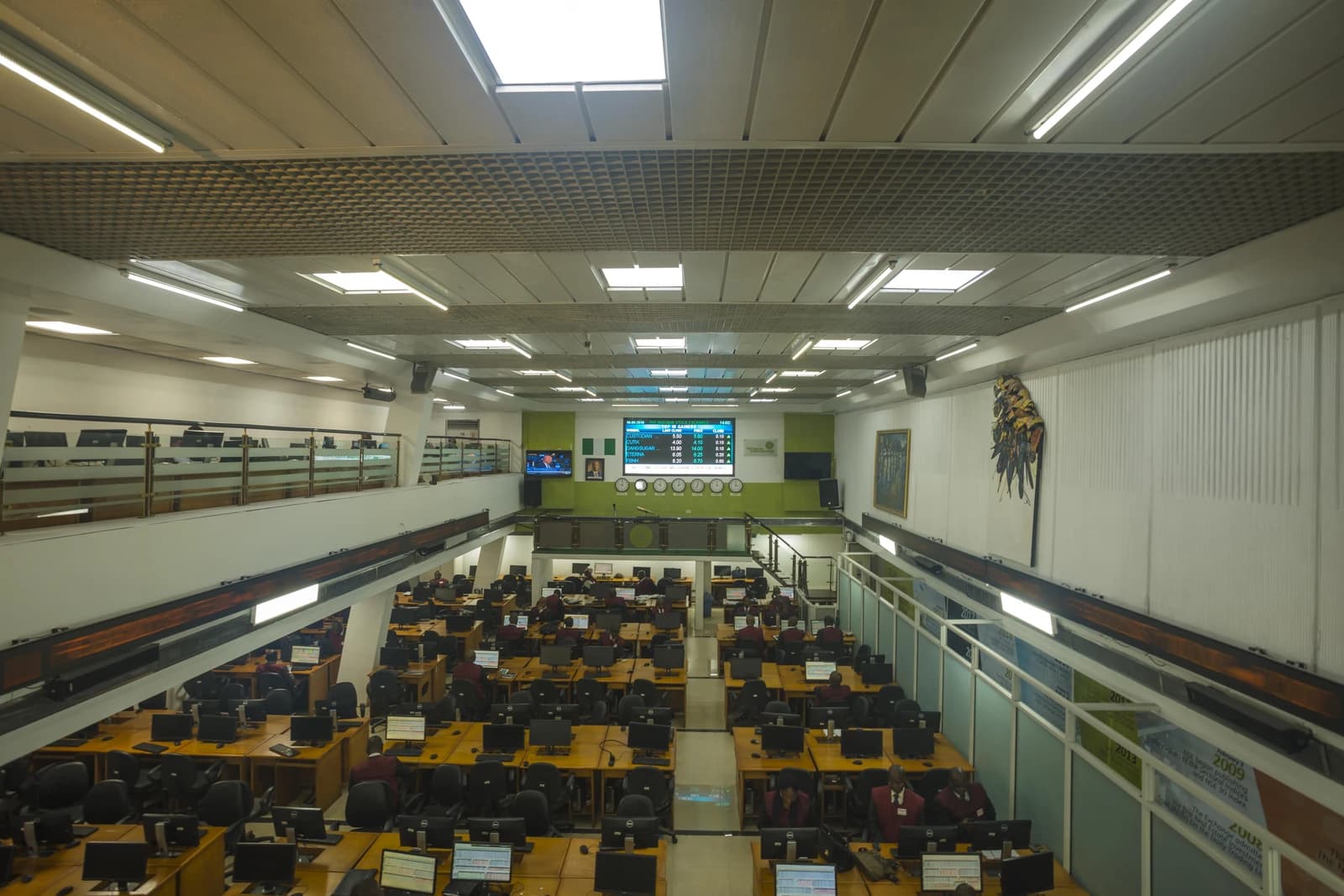 Overview of a stock exchange trading floor with traders at computers and a large display board.