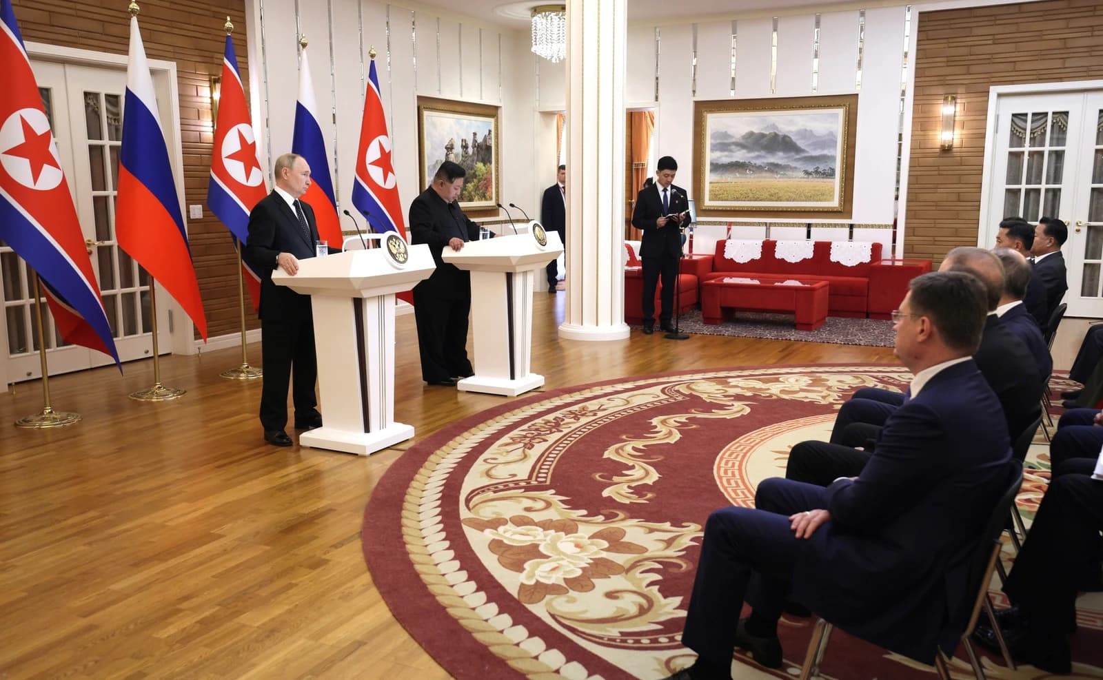 Vladimir Putin stands at a podium next to North Korean flags, facing forward.