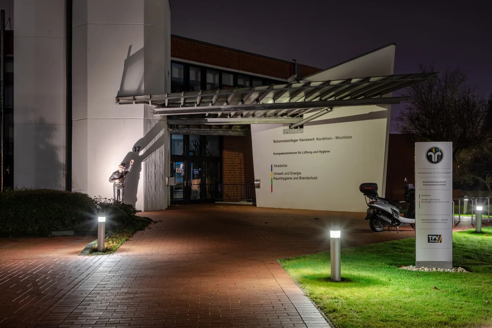 Exterior of Schornsteinfeger Handwerk Nordrhein-Westfalen, a chimney sweep vocational school, at night with a chimney sweep statue.
