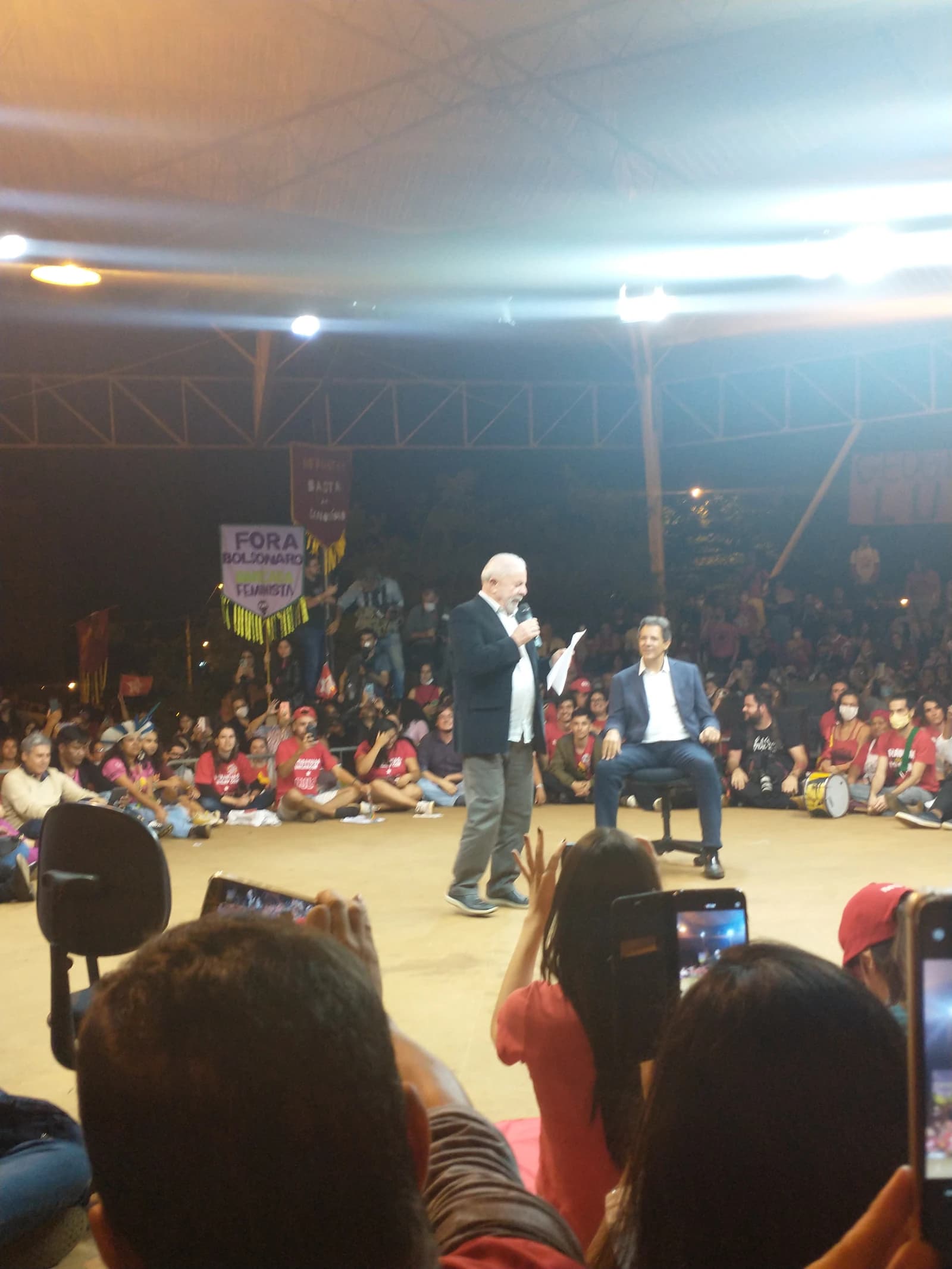 Luiz Inácio Lula da Silva speaking into a microphone with Fernando Haddad seated nearby.