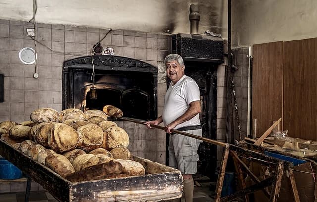 Baker using a peel to remove fresh bread loaves from a traditional oven.