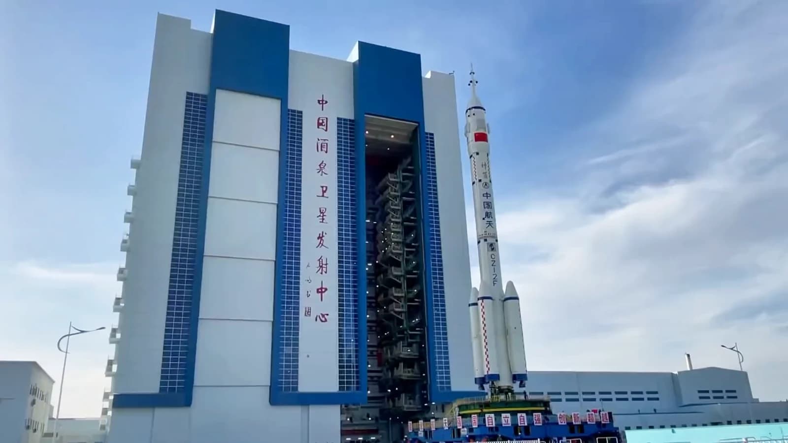 A large white and blue rocket stands next to a tall vertical assembly building at a launch site.