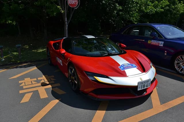 Red Ferrari SF90 Stradale with white racing stripes parked outdoors, a blue car is behind it.