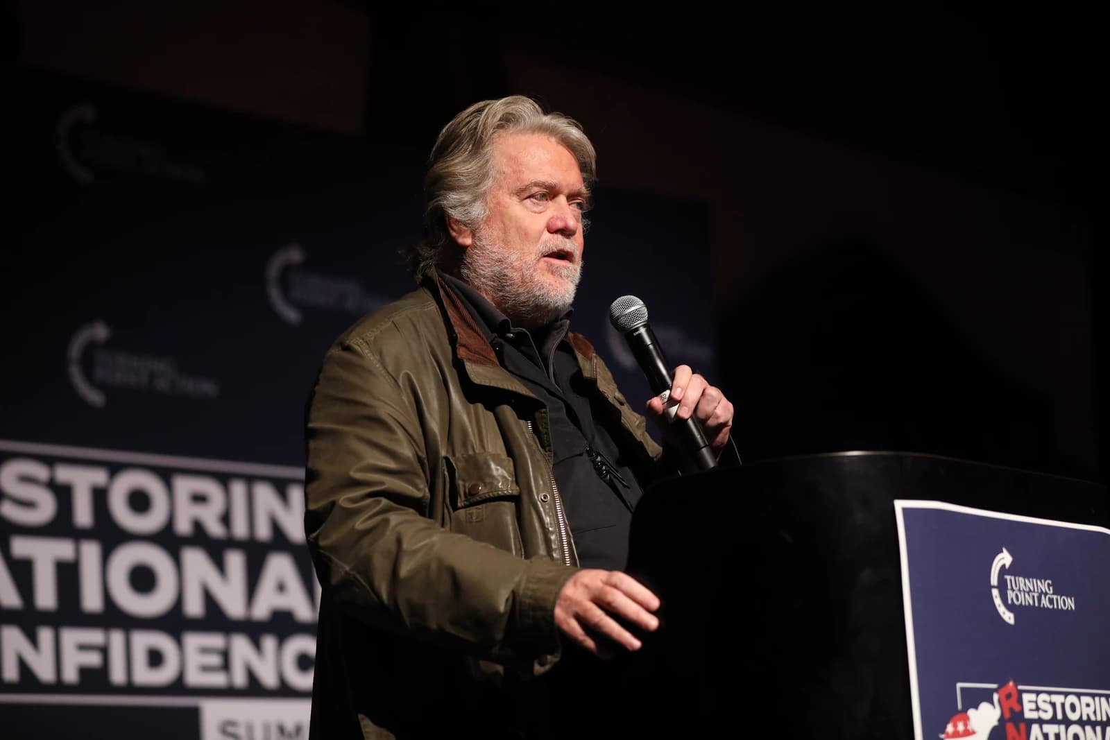 Steve Bannon speaking at a podium with a microphone in his right hand.