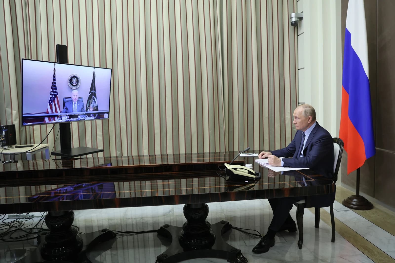 Vladimir Putin sitting at a large table, watching Joseph Biden on a screen.