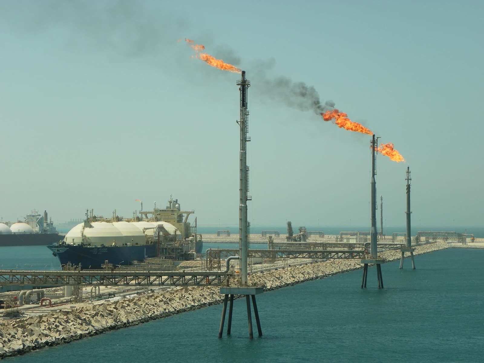 An LNG tanker docked at the Ras Laffan terminal with industrial flare stacks emitting flames.