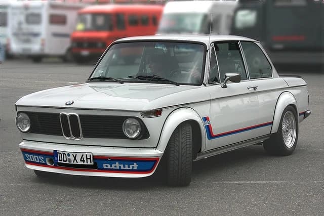 A white BMW 2002 Turbo with blue and red racing stripes parked on asphalt.