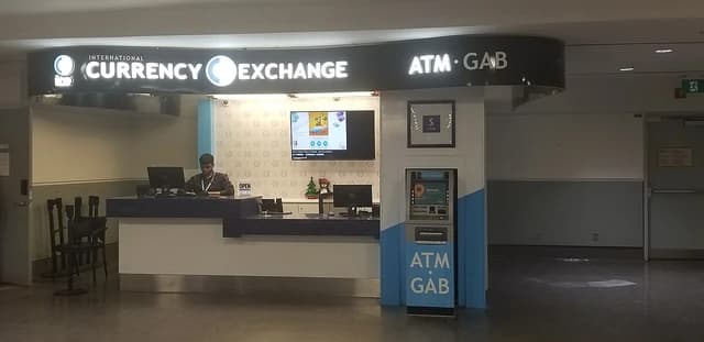 International Currency Exchange (ICE) office counter with an employee and ATM at an airport terminal.