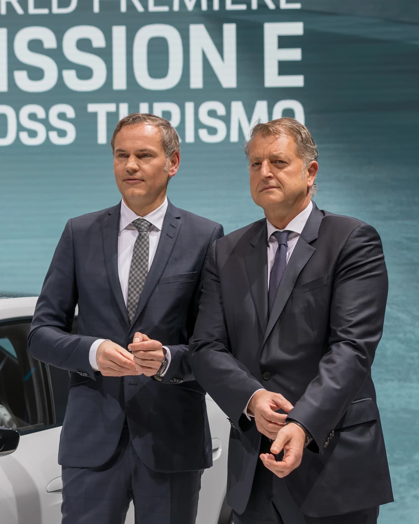 Two men in suits standing at a car show, with text on a screen behind them.